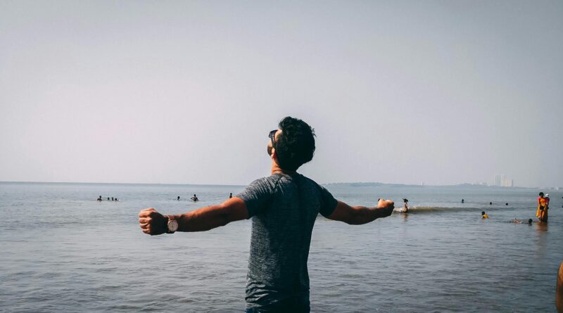 A man with open arms embracing freedom on a sunny beach day, surrounded by people enjoying the water.